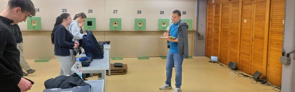 Serbian coach Goran Mikec standing in front of a group of young sport shooters, holding a copy of Shooting Notes, explaining the benefits of writing a shooting journal.