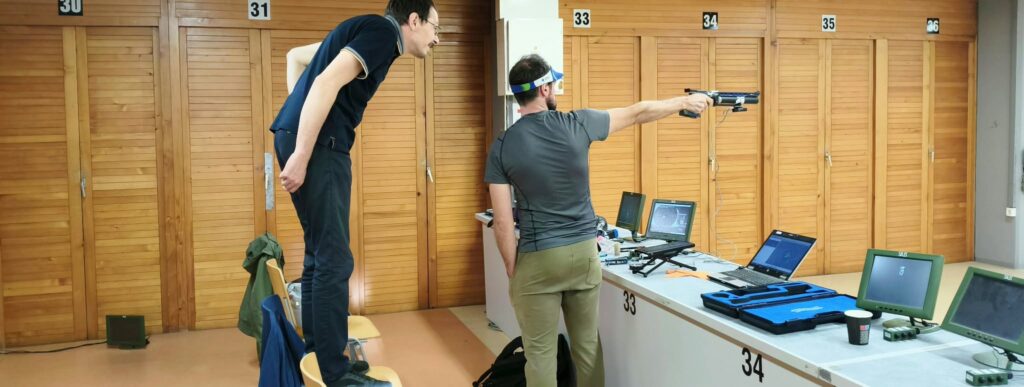 A pistol shooter aiming towards the target with his coach standing on a chair behind him and checking the movement of the pistol.